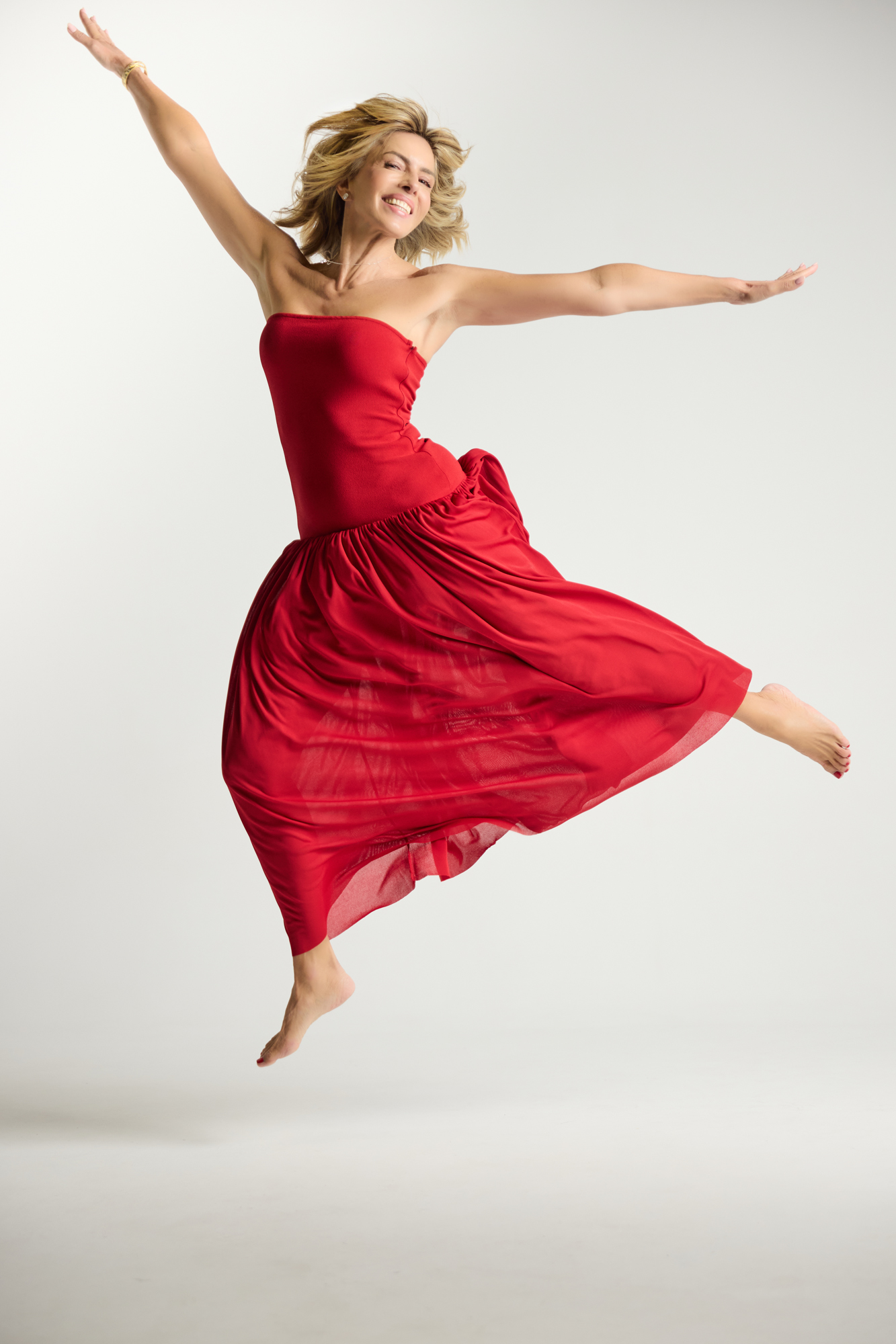 Esther Sarphatie jumping barefoot in a red strapless dress, smiling with arms outstretched in a studio.